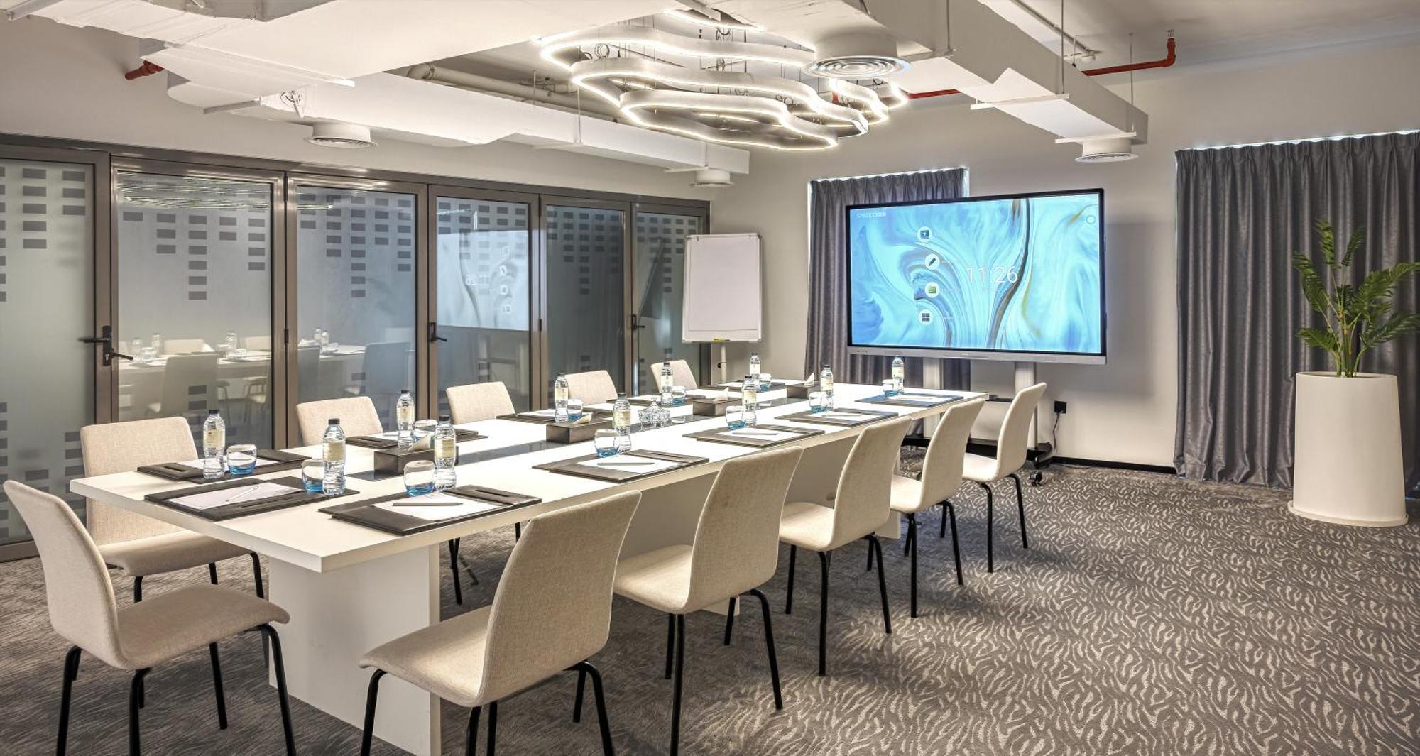 Aparthotel Millennium Atria Business Bay Dubái Exterior foto The photo shows a modern conference room set up for a meeting. There is a long table with several chairs arranged around it, each with a notepad and pen. A large screen displays a graphic or presentation, and there is a whiteboard in the corner. The 