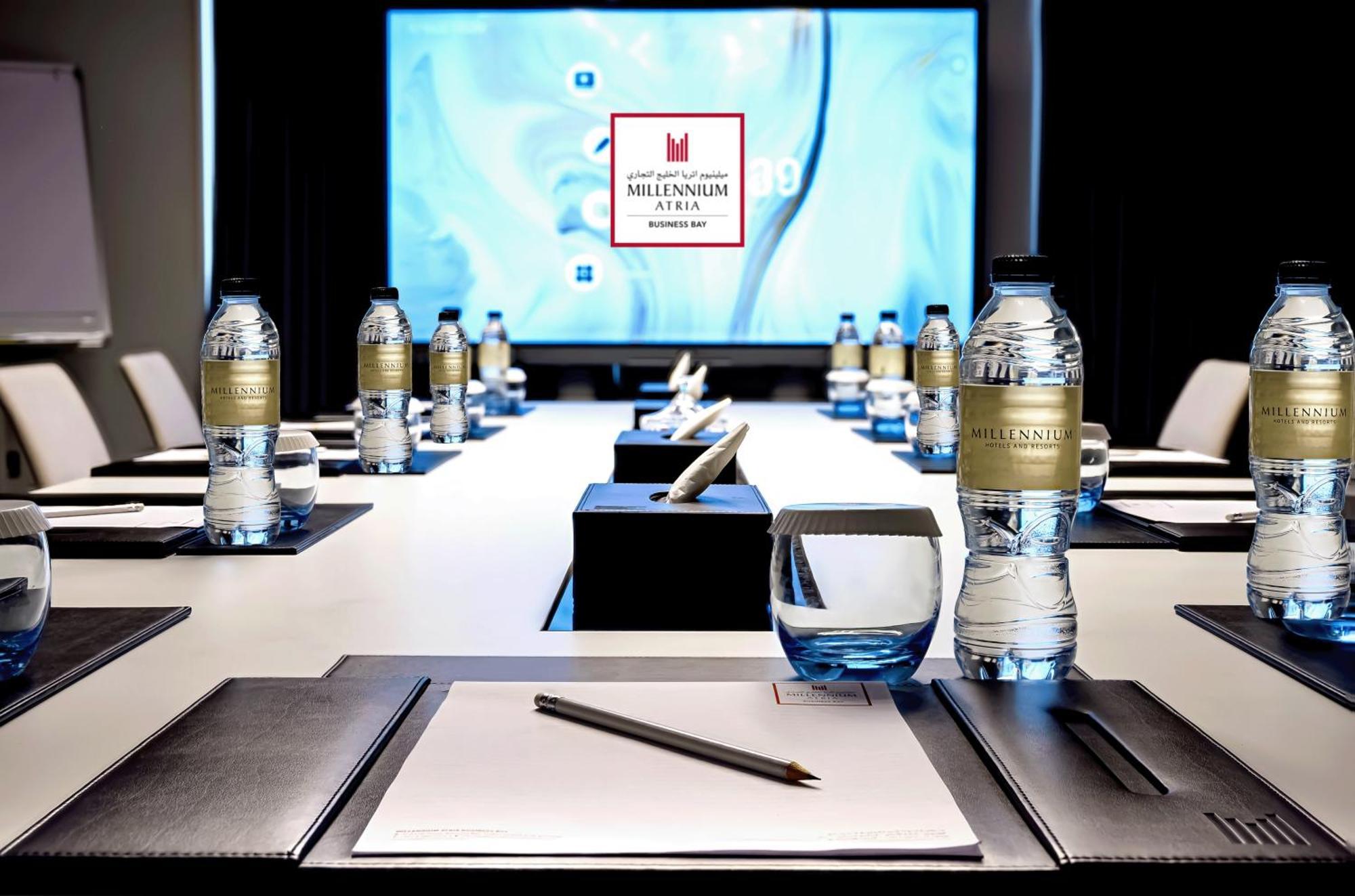 Aparthotel Millennium Atria Business Bay Dubái Exterior foto The photo shows a modern conference room setup. A long table is laid out with neatly arranged stationery, including notebooks and pens, at each seat. Several bottles of water are placed on the table. In the background, a large screen is displayed, li
