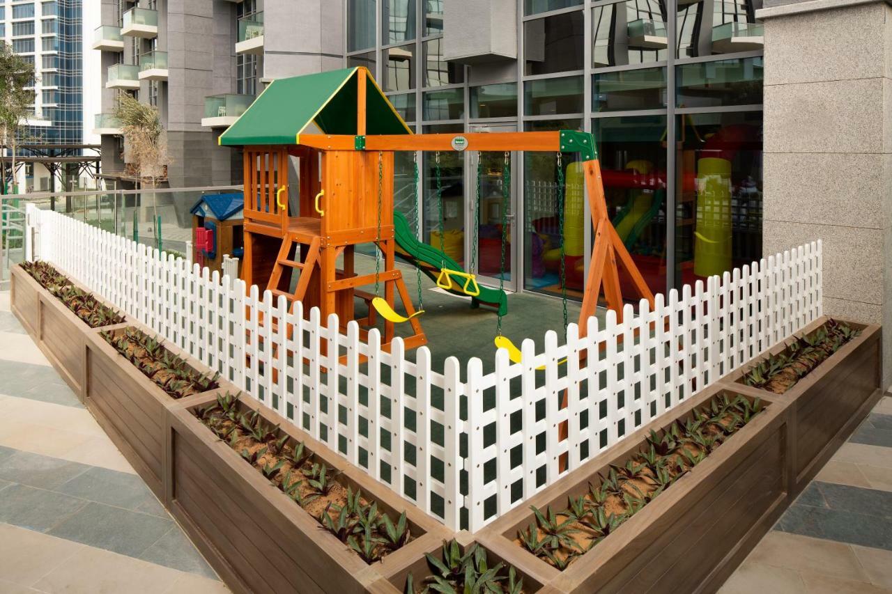 Aparthotel Millennium Atria Business Bay Dubái Exterior foto The image shows a children's playground area, which is enclosed by a white picket fence. The playground features a wooden play structure with a slide, swings, and climbing elements. Surrounding the play area are planters with greenery, and the backdr