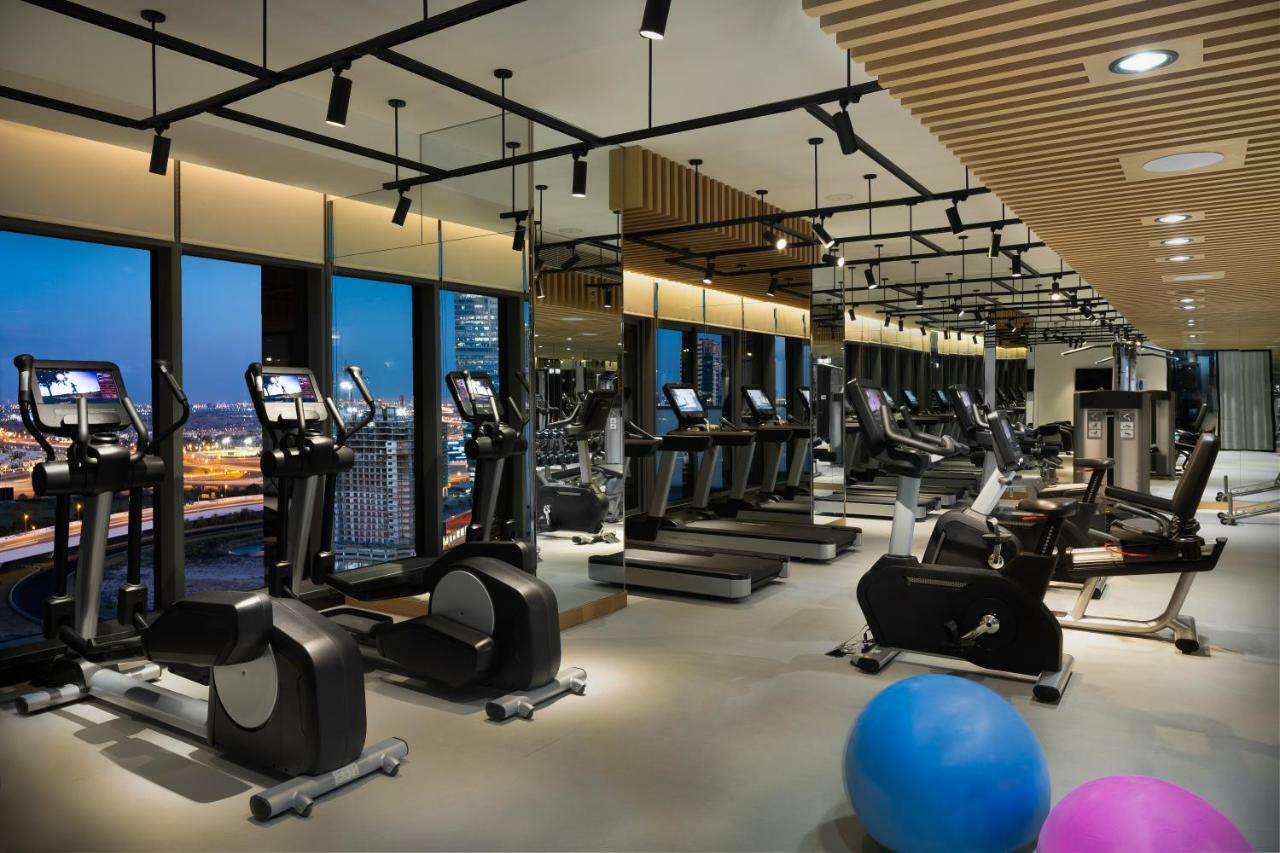 Aparthotel Millennium Atria Business Bay Dubái Exterior foto The photo shows a modern gym with various types of exercise equipment available. There are several treadmills and elliptical machines arranged in rows, with large windows in the background providing a view of the city. The lighting is bright and cont