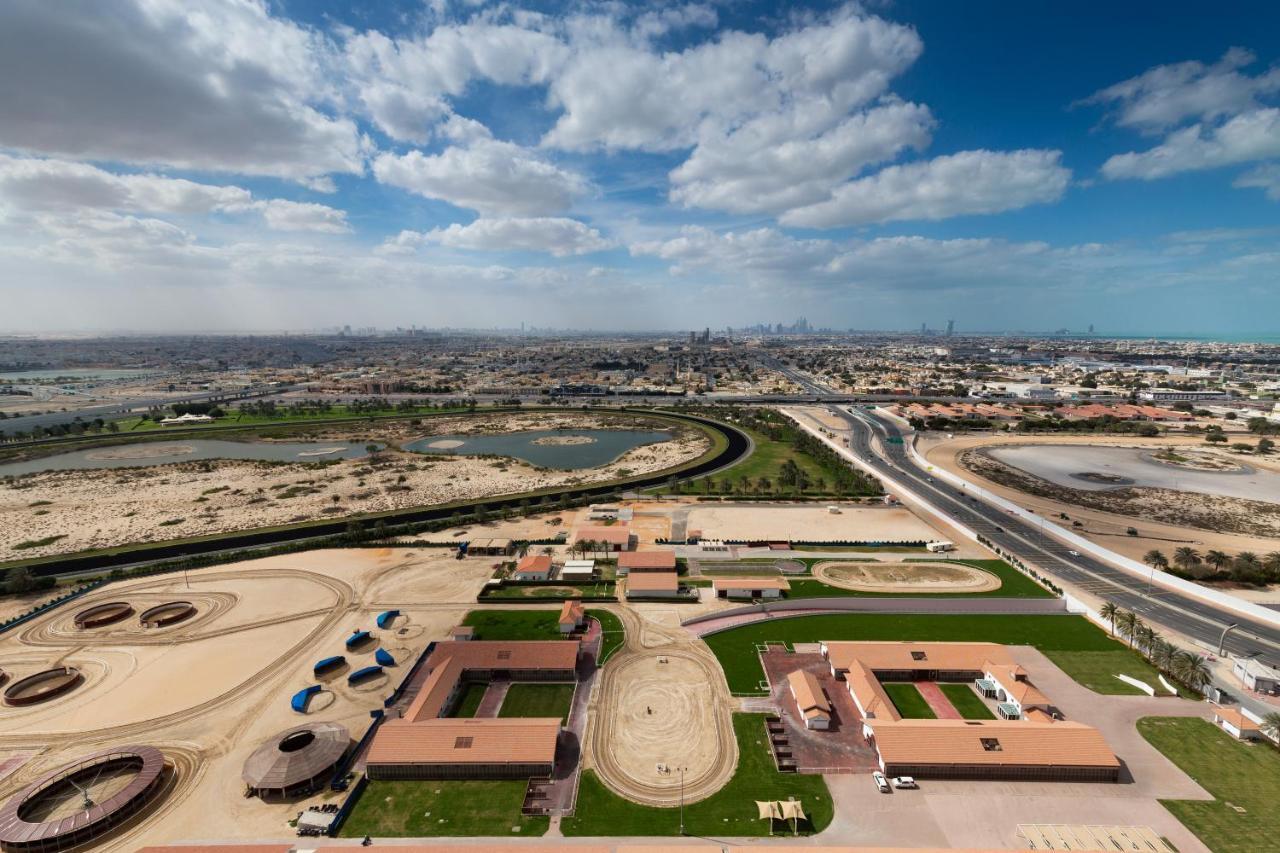 Aparthotel Millennium Atria Business Bay Dubái Exterior foto The photo shows a panoramic view of a large landscape with expansive open areas, buildings, and a curved track. In the foreground, there are several stable structures and facilities associated with horse training or racing, surrounded by well-maintai
