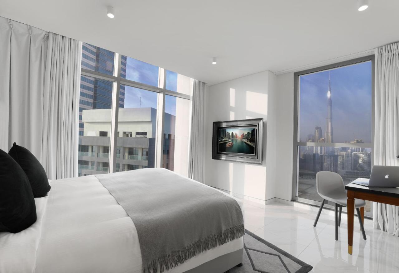 Aparthotel Millennium Atria Business Bay Dubái Exterior foto The photo depicts a modern hotel room that features a large bed with a gray throw blanket. There are large windows allowing natural light to fill the space, showcasing a city skyline view. A flat-screen television is mounted on the wall opposite the 